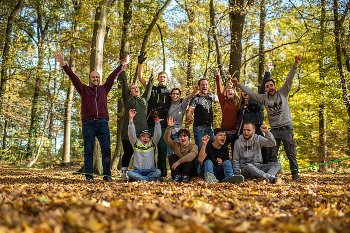 eine Gruppe von Schüler:innen und Lehrer:innen steht im Herbstwald. Sie jubeln in die Kamera und sind sehr fröhlich!